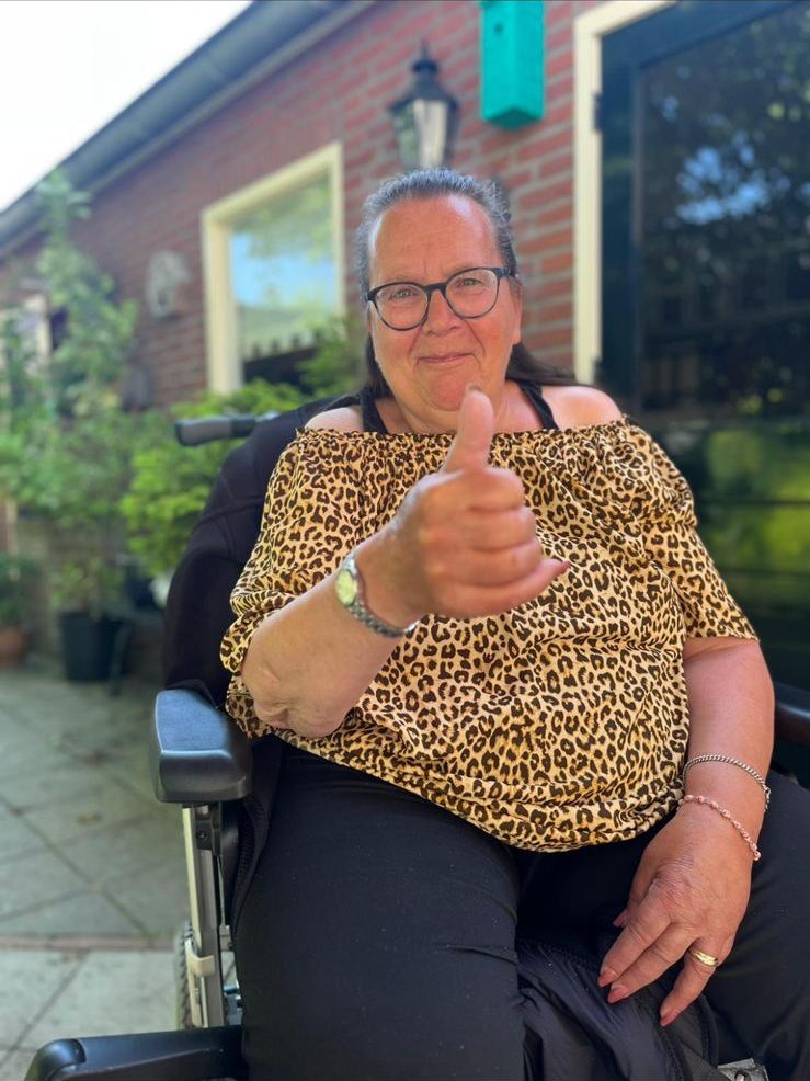 Marian in haar rolstoel voor haar huis, lachend met haar duim omhoog